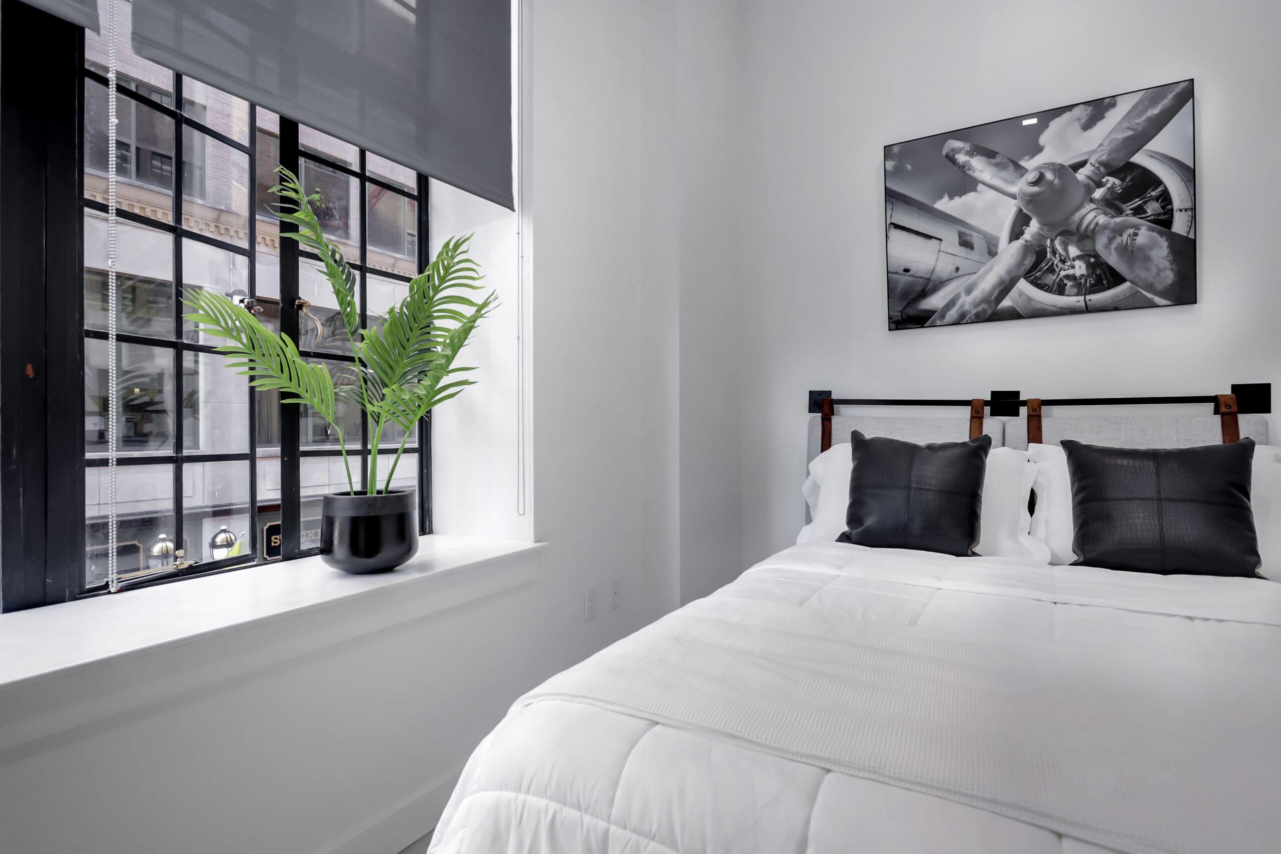 Bedroom at 99 Pratt Street Apartment bedroom with white bedding, black pillows, industrial windows, and a potted plant