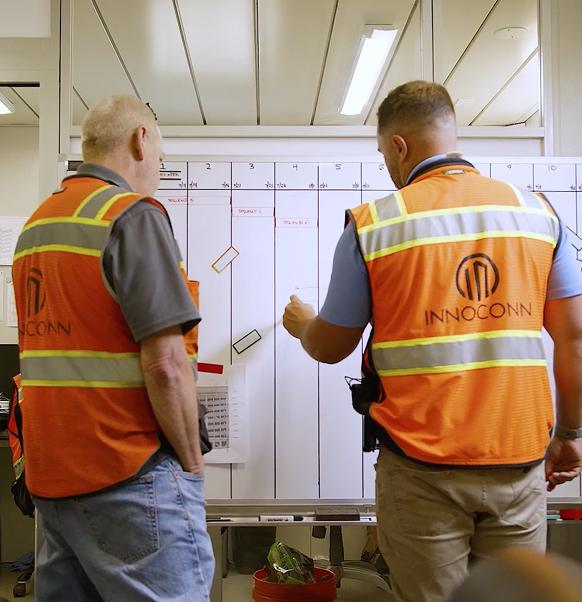 Two construction workers in orange high-visibility vests reviewing plans posted on a wall
