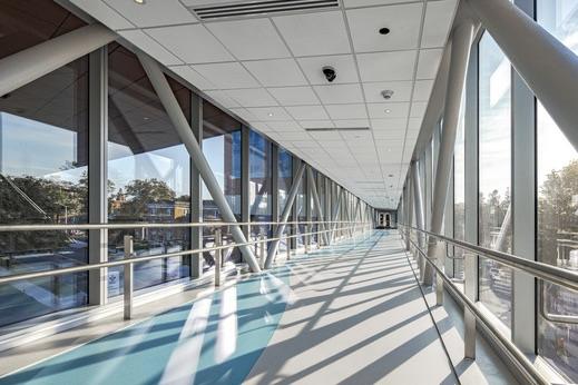 Walkway at LPRI CCMC Garage Interior of a sunlit glass-enclosed pedestrian skybridge with diagonal steel supports and floor-to-ceiling windows
